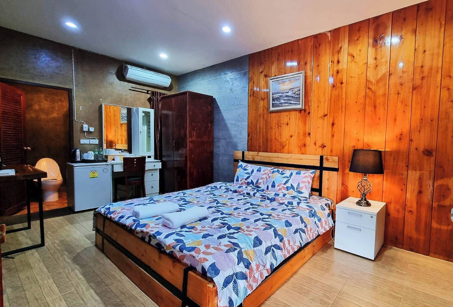Een slaapkamer met een houten hoofdeinde, een bed met een bedovertrek met patroon, een nachtkastje met een lamp, een klein bureau met een stoel, een kledingkast en een airconditioning.