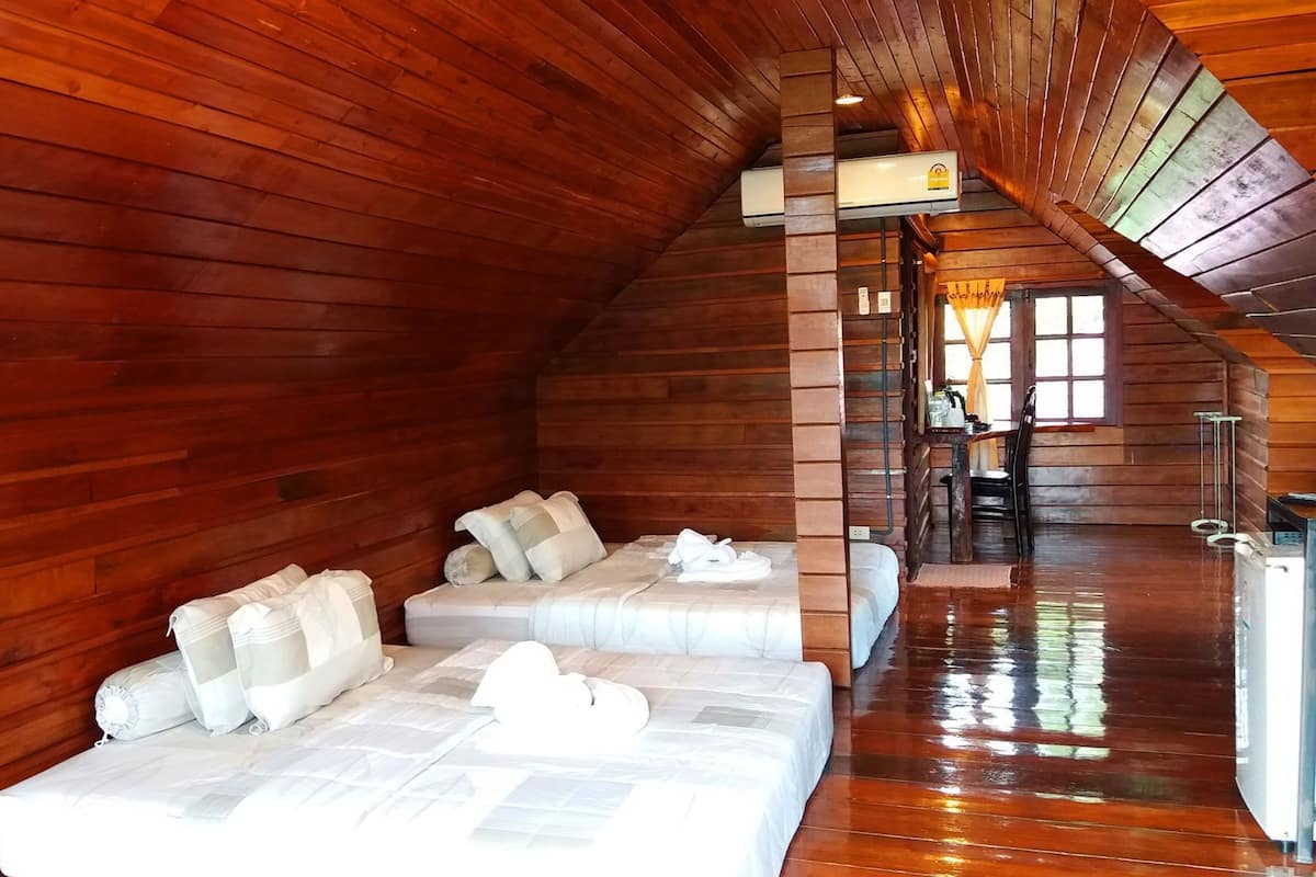 A log cabin interior with a loft bedroom featuring two beds, a wooden ceiling, and a window.