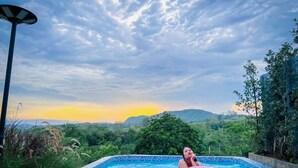 A swimming pool with a person on a pink float, surrounded by lush greenery and a mountainous backdrop. - Kirirath Resort (Pak Chong)