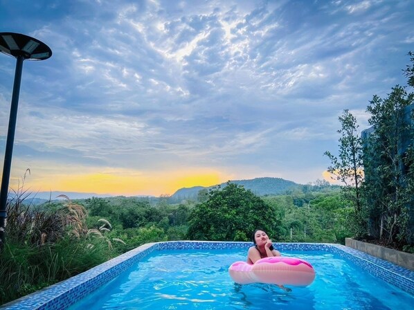 A swimming pool with a person on a pink float, surrounded by lush greenery and a mountainous backdrop. - Kirirath Resort (Pak Chong)