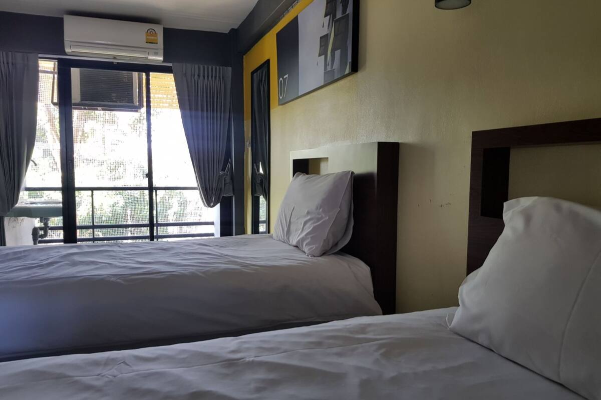 A hotel room with two beds, a large window with curtains, an air conditioning unit, and a wall-mounted picture.