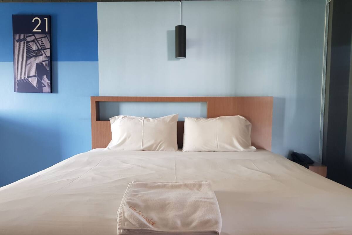 A neatly made bed with a folded towel, a wooden headboard, and a wall-mounted number sign.