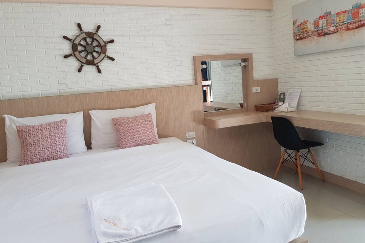 A hotel room with a bed, a desk, a chair, a ship's wheel decoration, and a painting on the wall.