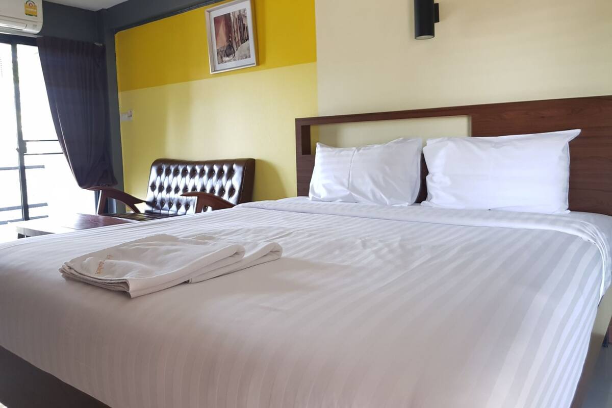 A neatly made bed with white linens and a folded towel, a wooden headboard, a chair, and a wall-mounted picture.