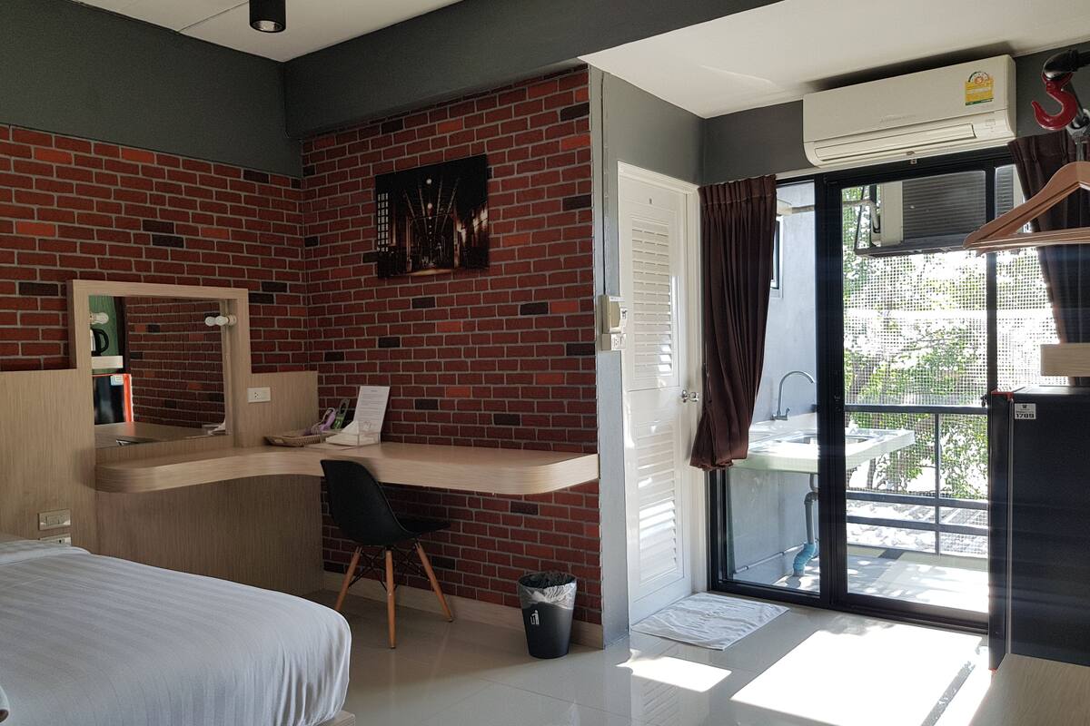 A hotel room with a brick wall, a desk with a chair, a bed, a sliding glass door, and an air conditioning unit.