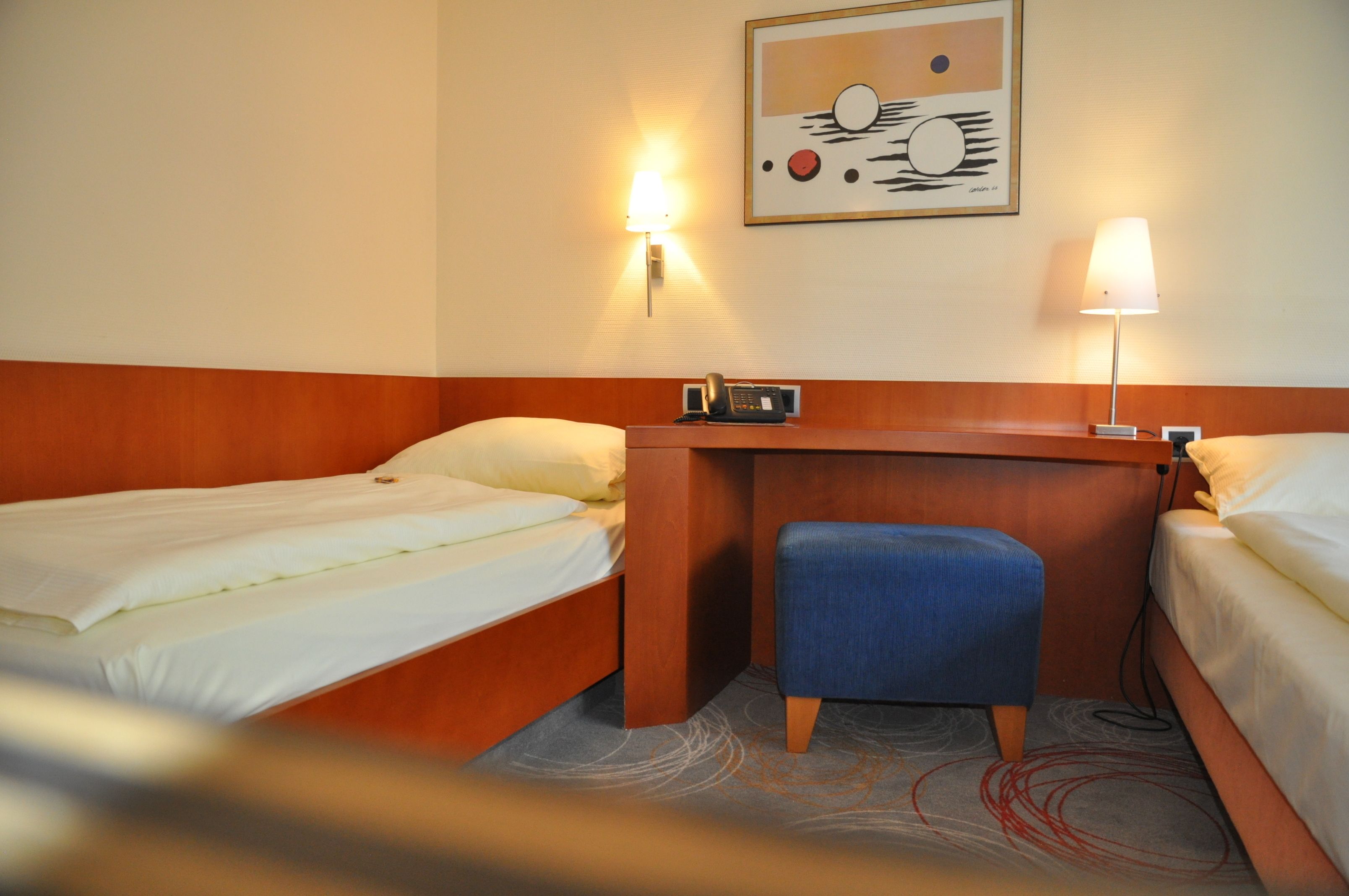 Een hotelkamer met twee eenpersoonsbedden, een houten bureau, een blauwe stoel en een wandlamp.