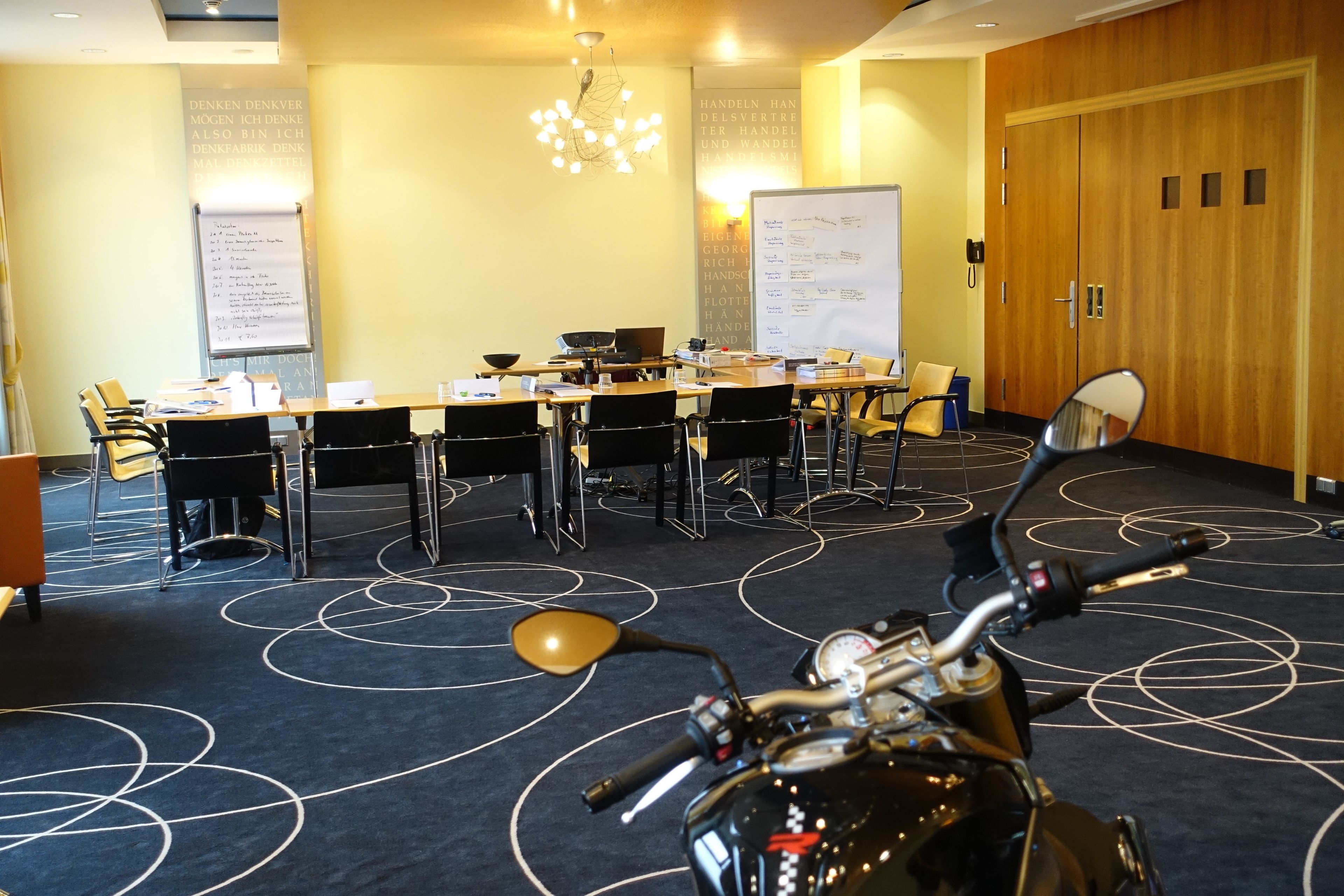Une salle de conférence avec une longue table, des chaises, un tableau blanc et une moto au premier plan.