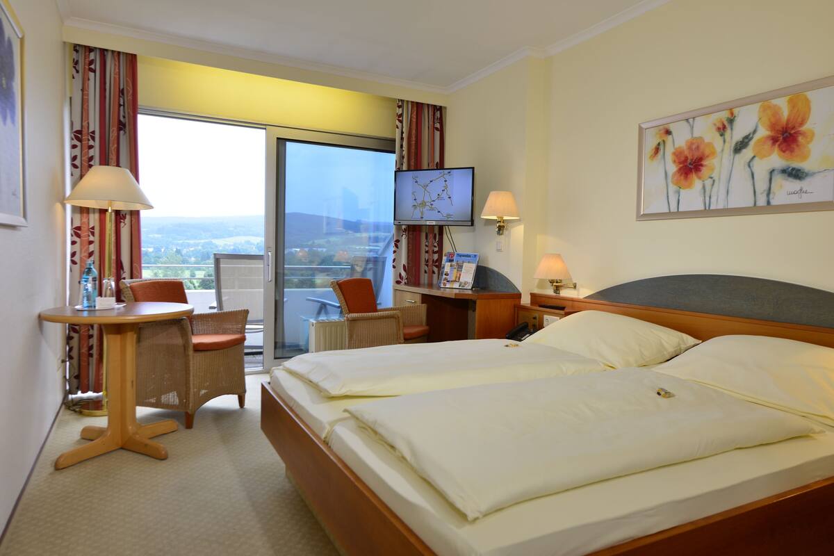 Habitación de hotel con cama, escritorio, silla, televisión y balcón con vistas.