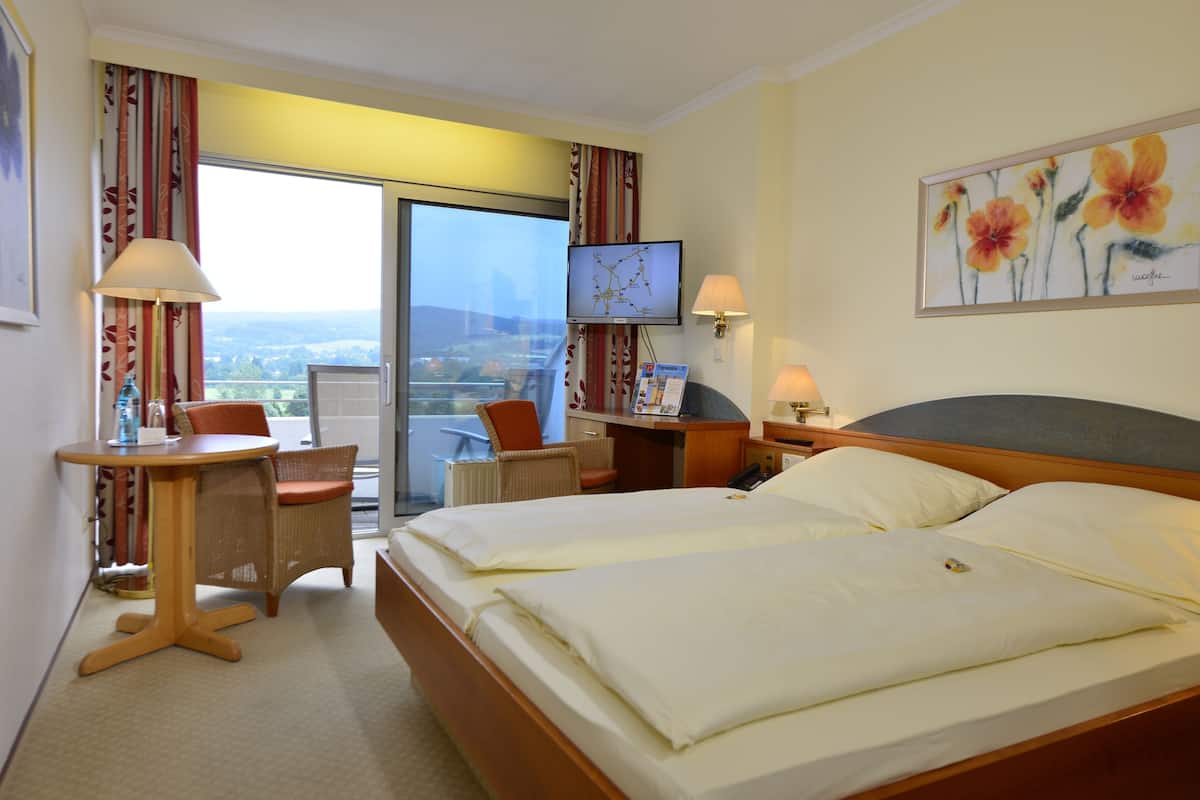Habitación de hotel con cama, escritorio, silla, televisor y balcón con vistas.