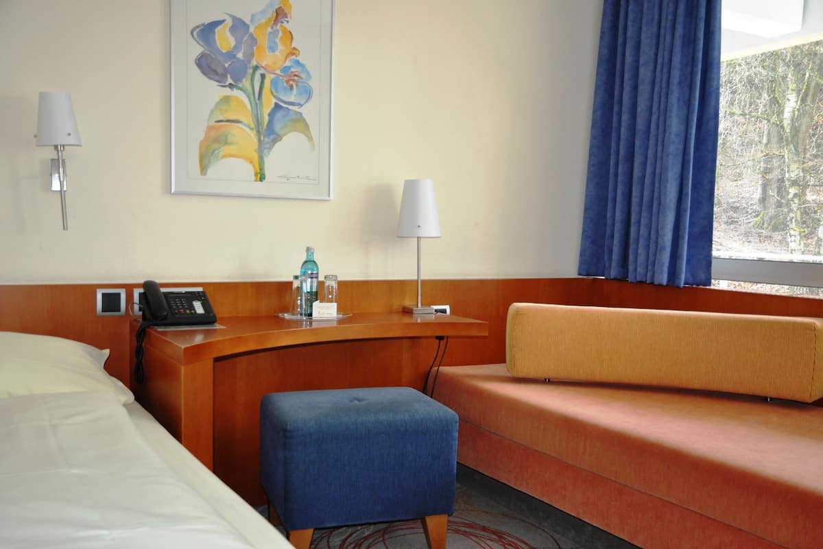 Una habitación de hotel con cama, escritorio con teléfono, lámpara, sillón y ventana con cortinas.