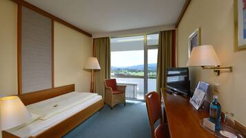 A hotel room with a bed, a chair, a desk with a TV, a glass of water, and a view of the outdoors.