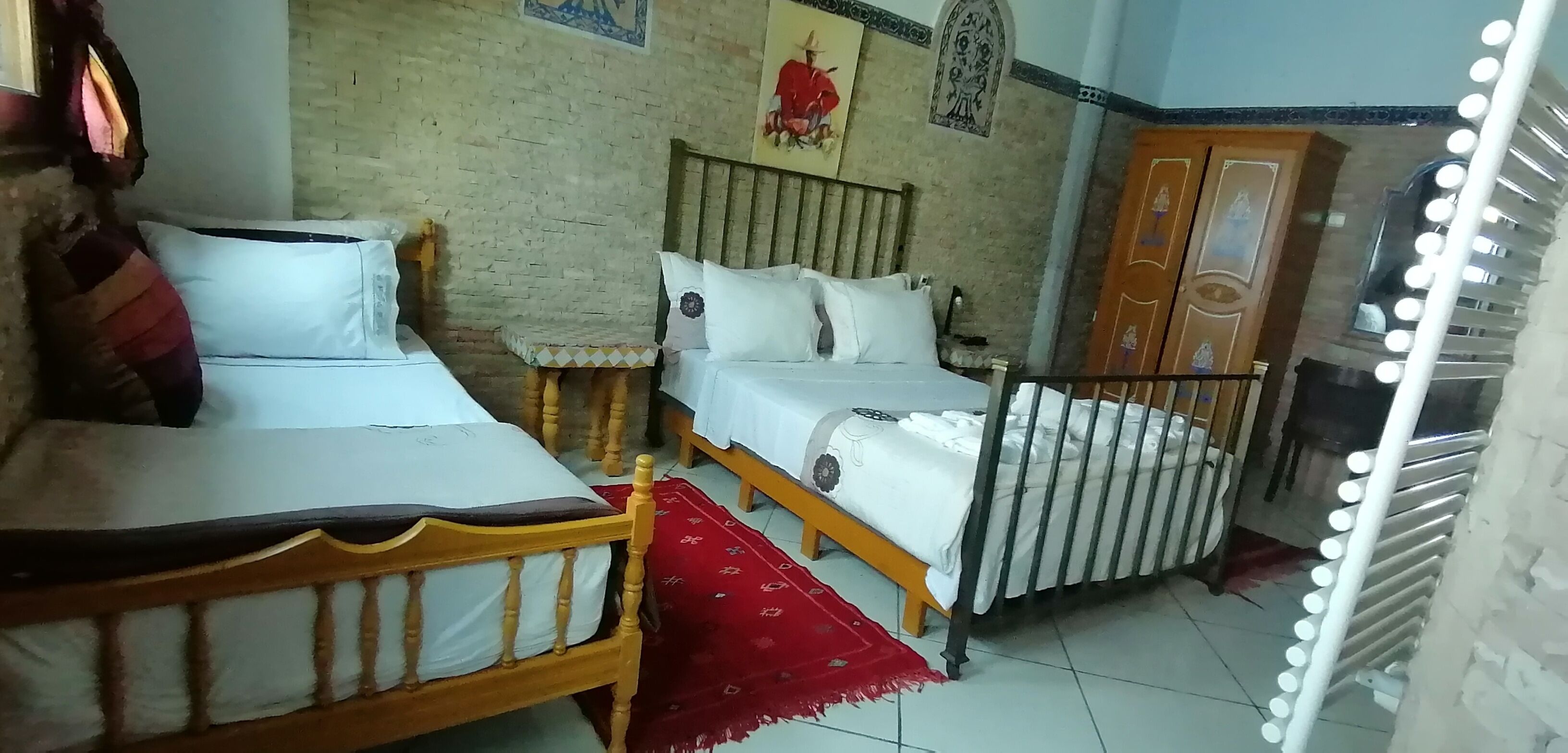 Een kamer met twee houten bedden, een tapijt met een rood patroon, een met decoraties beklede bakstenen muur en een spiegel met verlichting.