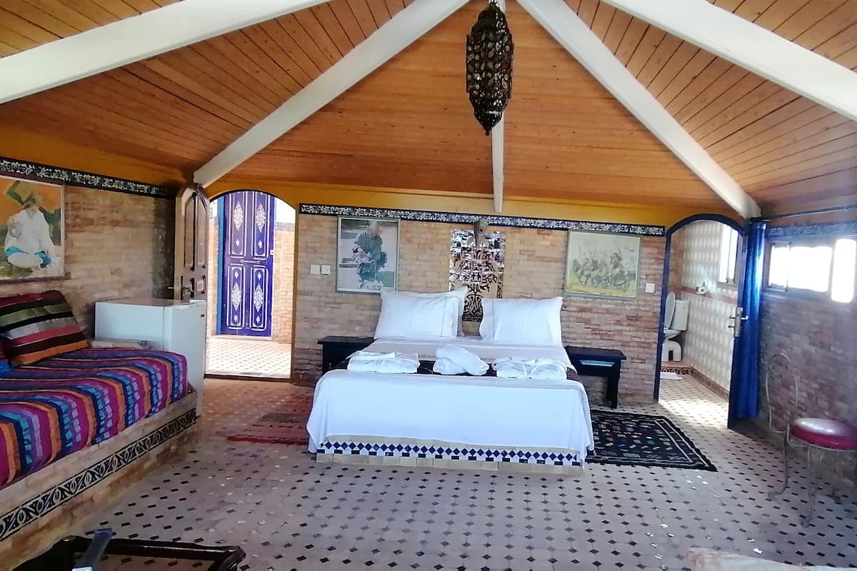 Una habitación con techo de madera alta, una cama con ropa blanca, un sofá colorido y un armario azul.