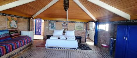 Une pièce avec un haut plafond en bois, un lit recouvert de draps blancs, un canapé coloré et une armoire bleue.