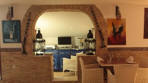 Un comedor con sillas de mimbre blancas, una mesa dispuesta con platos y vasos, y una vista a través de un arco hacia una cocina con armarios azules y un televisor.