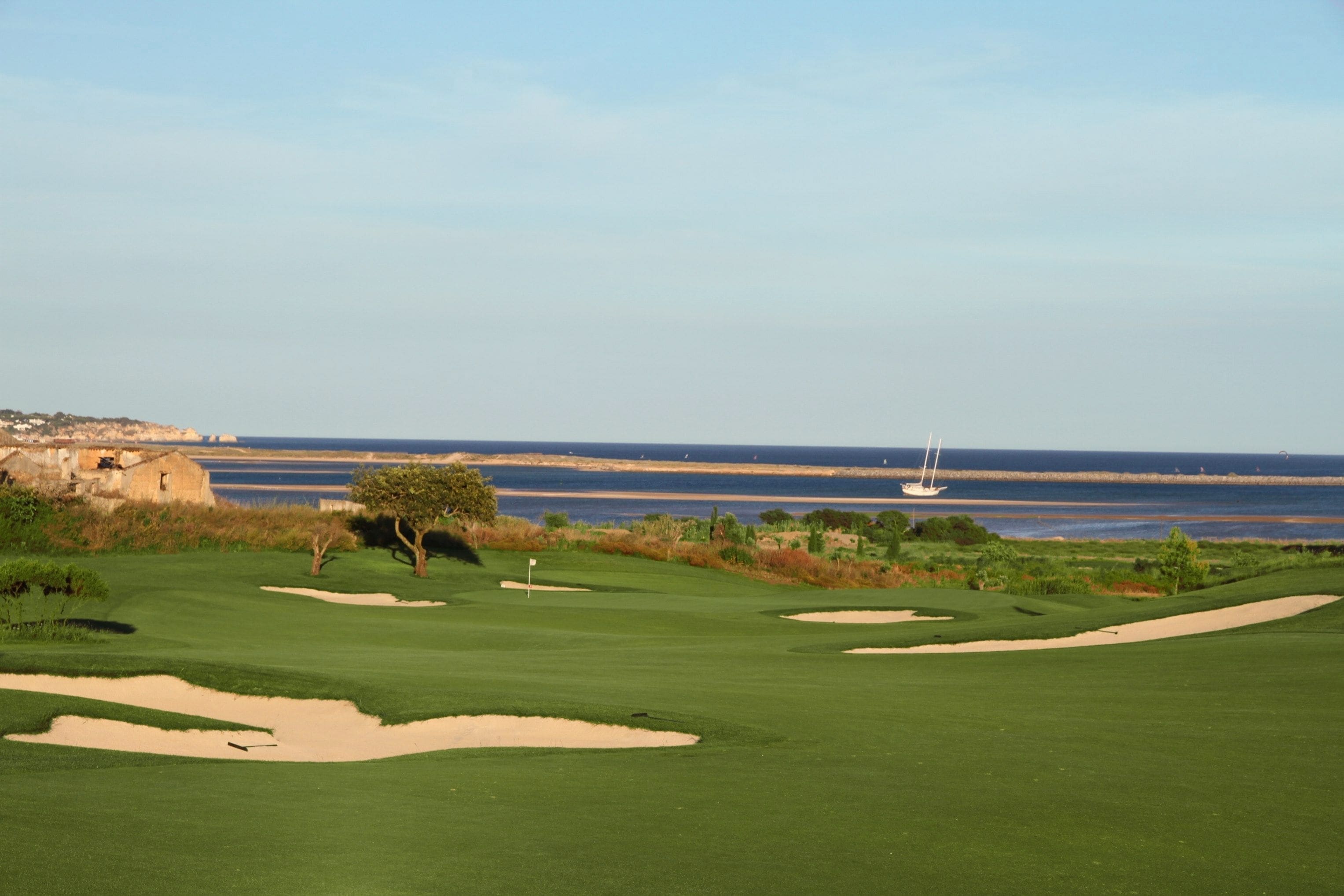 Un campo de golf con búnkeres y un mástil de bandera, con vistas a una zona costera con una playa de arena y un velero a lo lejos.
