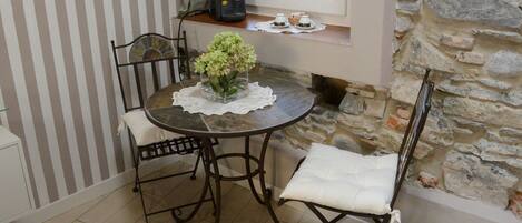 A dining area with a round table, two chairs, a vase with flowers, and a window with a stone wall.