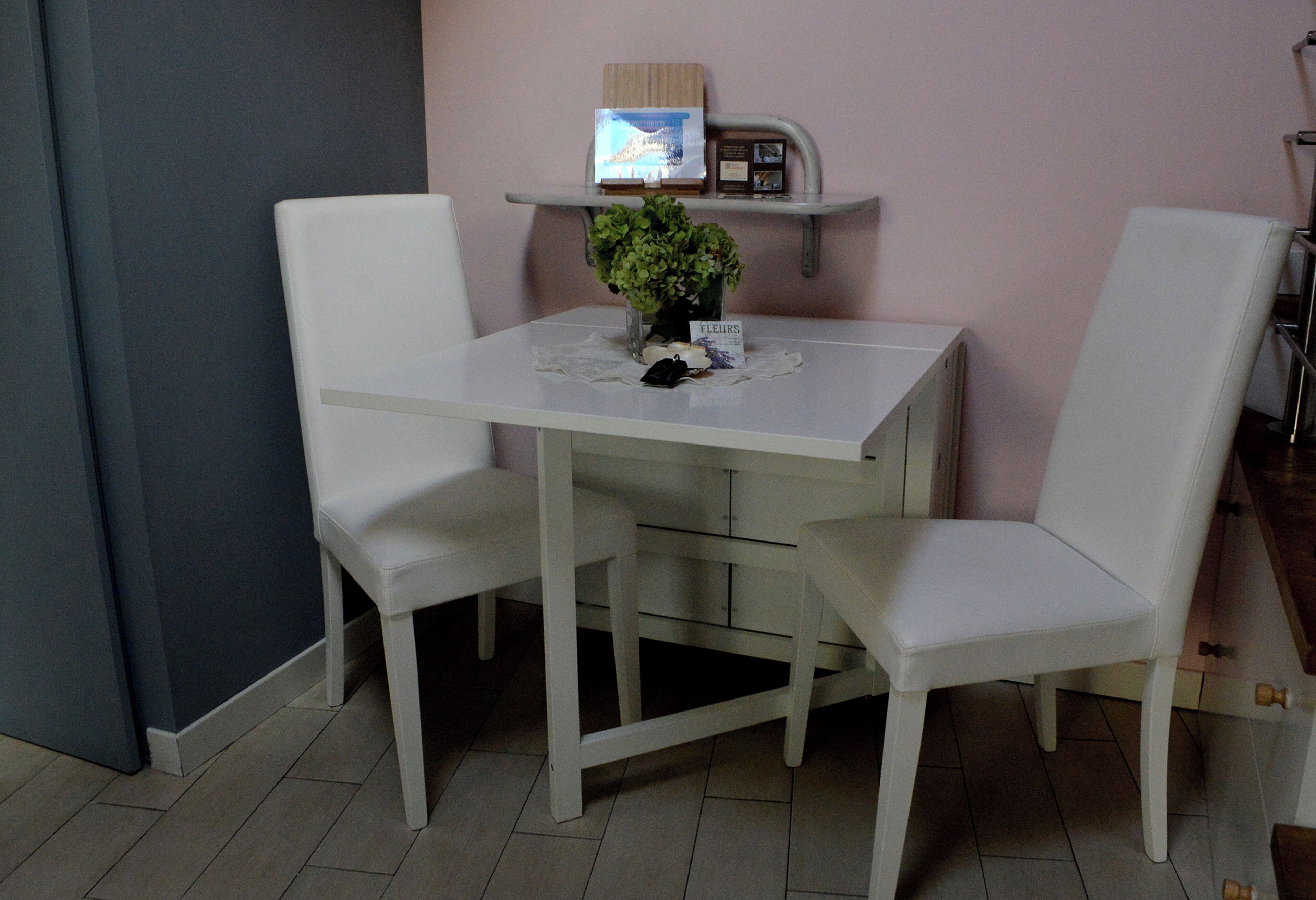 A white dining table with two chairs, a small potted plant, and a shelf with a picture and some items.