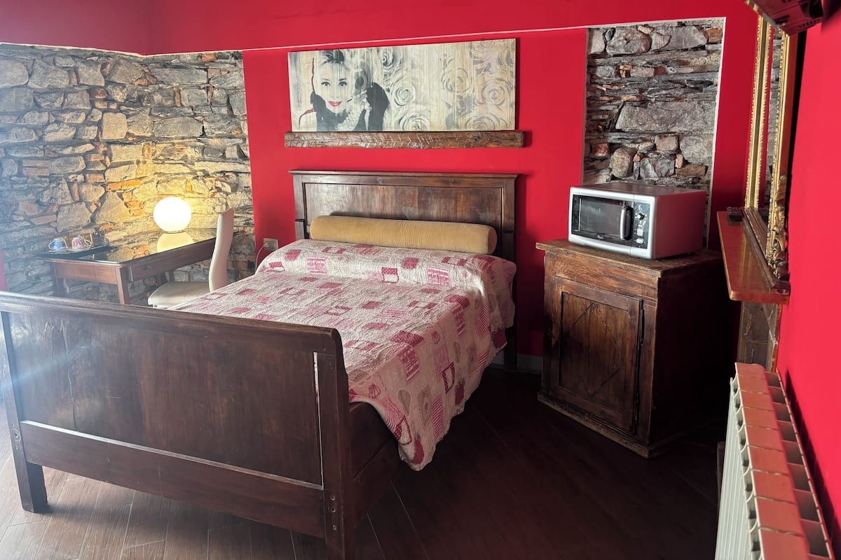 A bedroom with a wooden bed, a stone wall, a red wall, a painting, a microwave, and a cabinet.