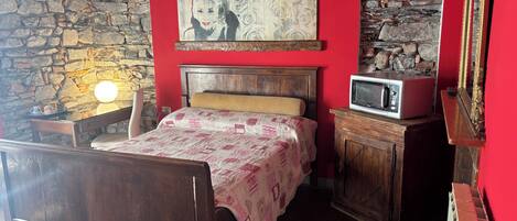 A bedroom with a wooden bed, a stone wall, a red wall, a painting, a microwave, and a cabinet.