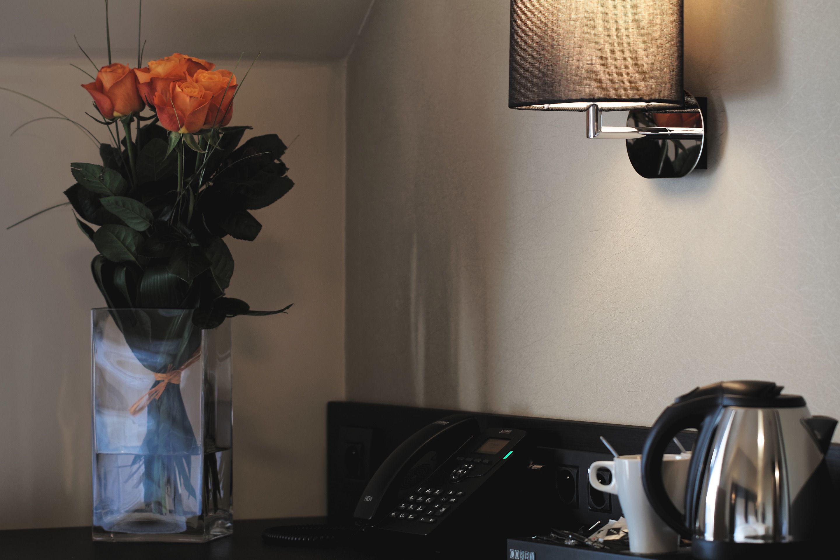 Eine transparente Glasvase mit Blumen auf einem Tisch, ein an der Wand befestigtes Telefon und ein Wasserkocher.
