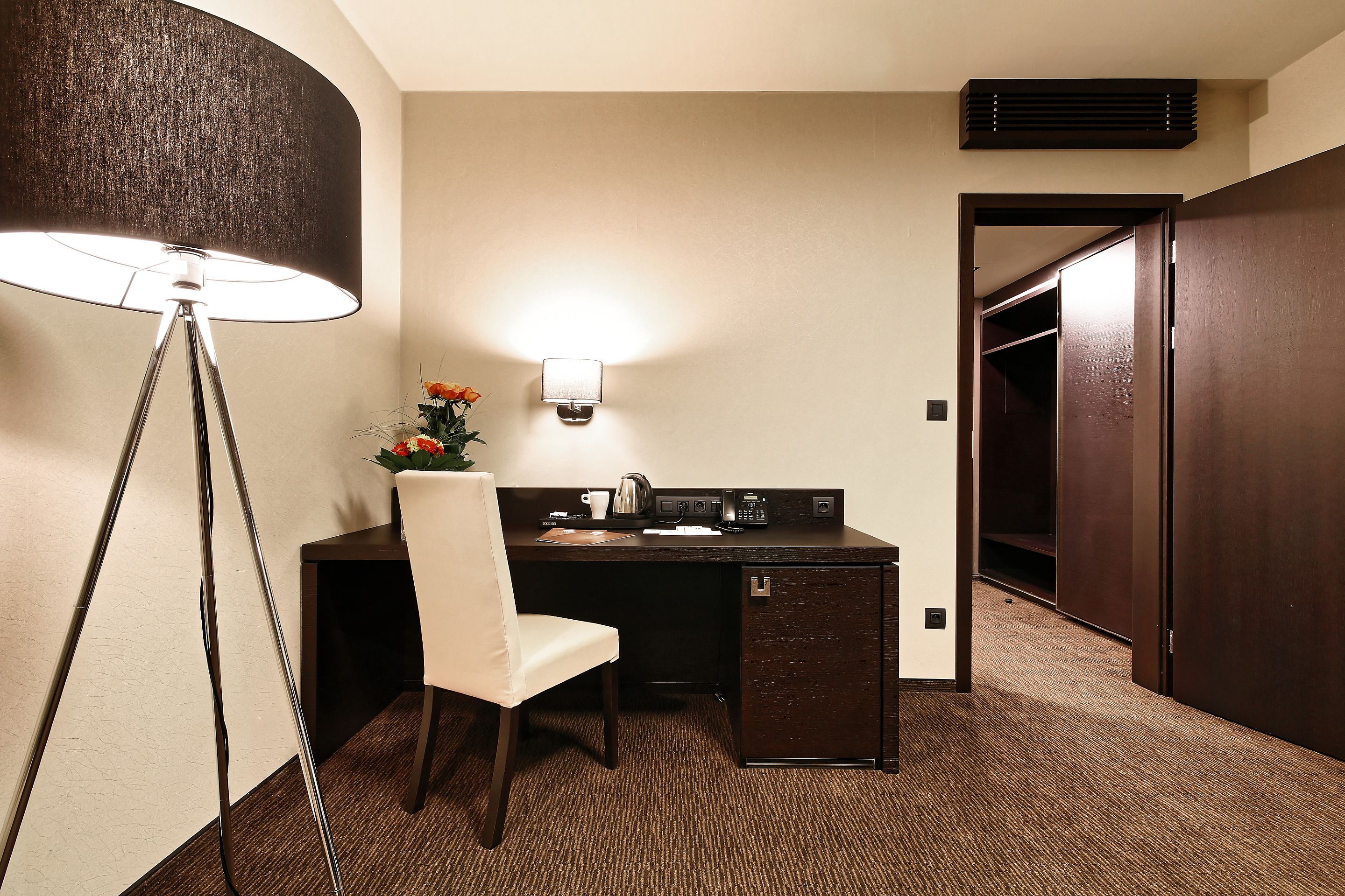 A hotel room with a desk, chair, lamp, and a small kitchenette.