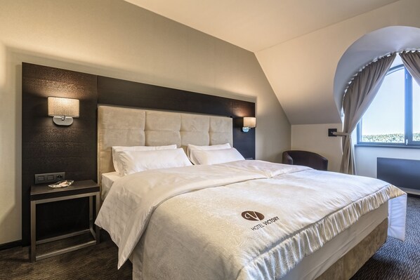 A hotel room with a large bed, two bedside tables with lamps, a small table with a lamp, and a view of the outdoors through a window with curtains.
