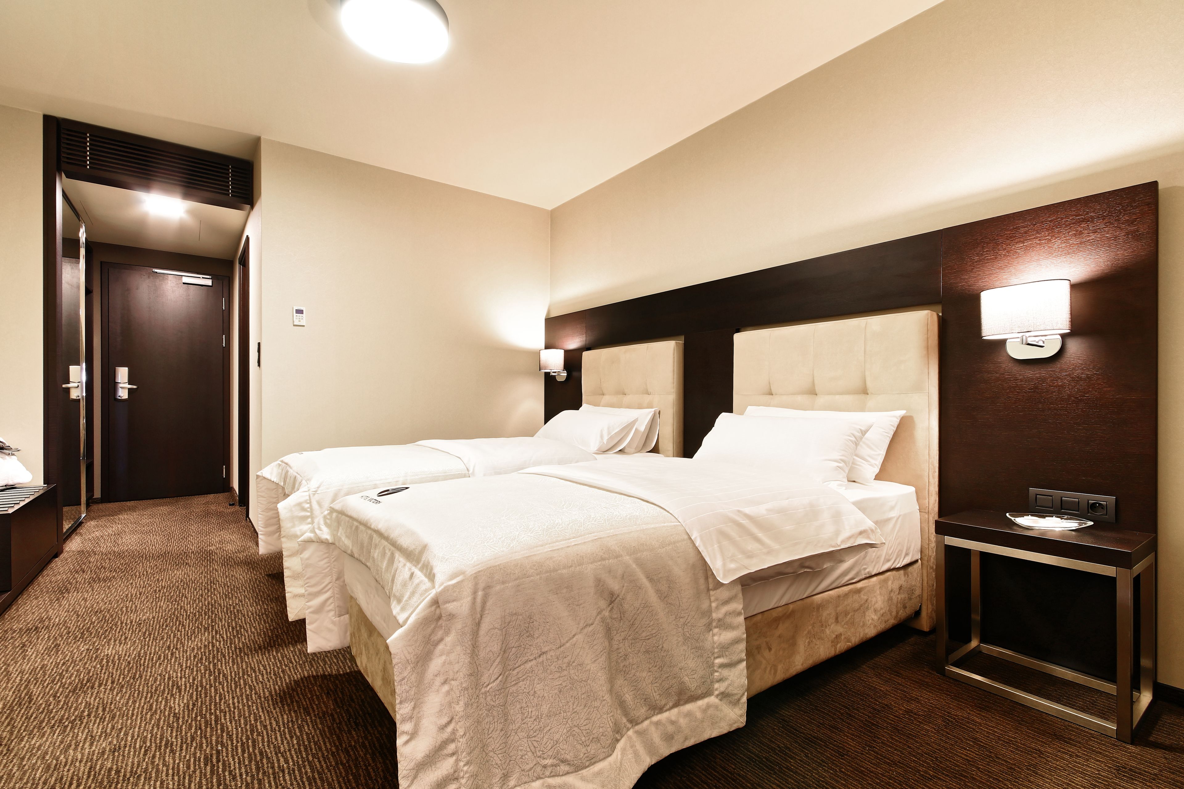 A hotel room with two beds, a bedside table, a nightstand, and a wall-mounted light fixture.