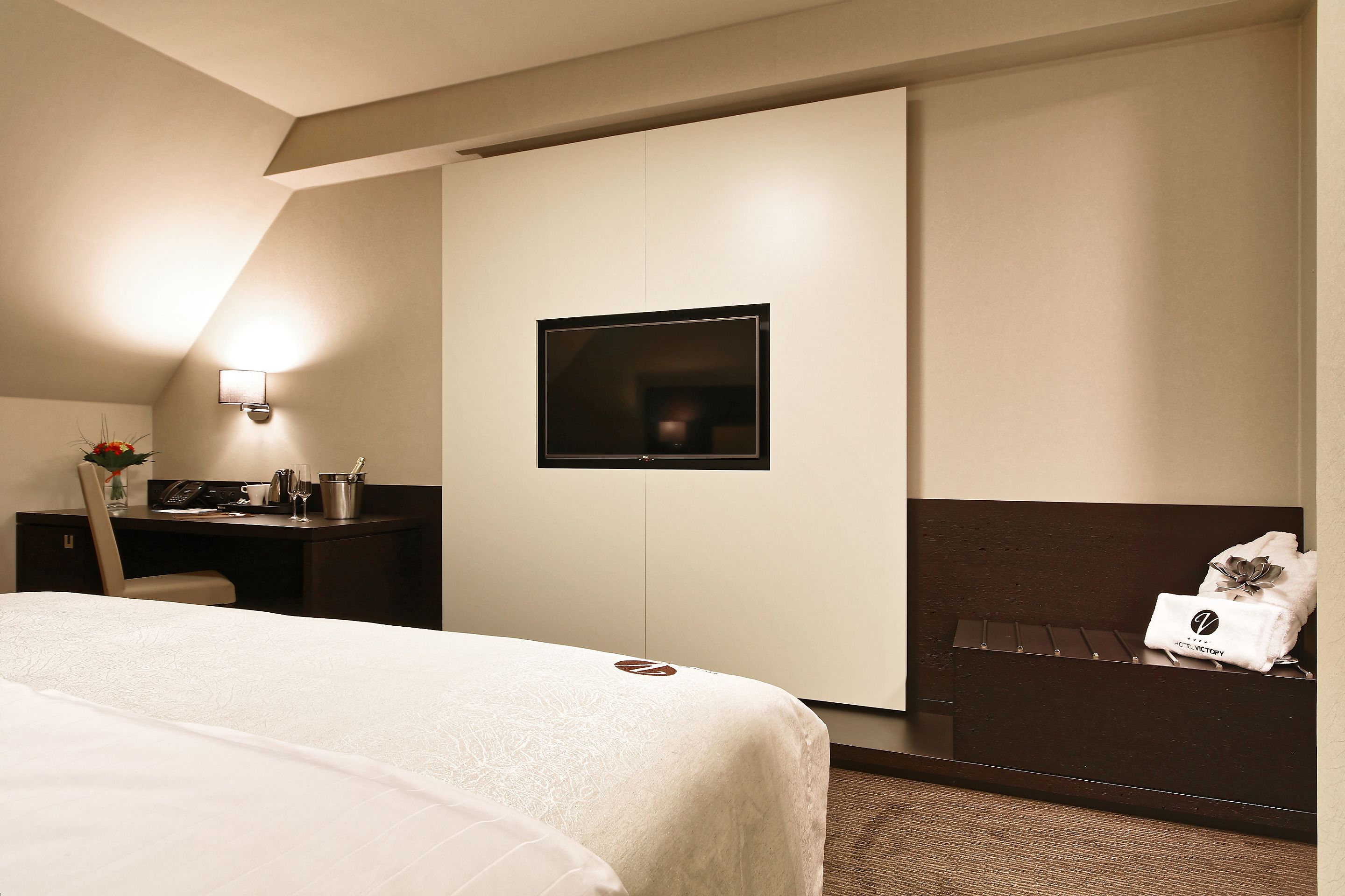Habitación de hotel con cama, escritorio con lámpara, televisión y un pequeño rincón de descanso con toalla.