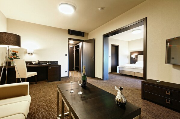 Habitación de hotel con escritorio, dos sillas, cama, cómoda, espejo y una botella de champán sobre la mesa.