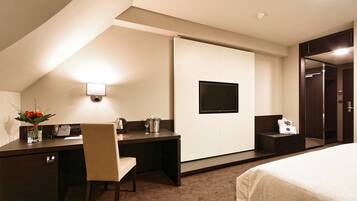 A hotel room with a bed, a desk with a chair, a small table with a vase of flowers, a telephone, and a television mounted on the wall.
