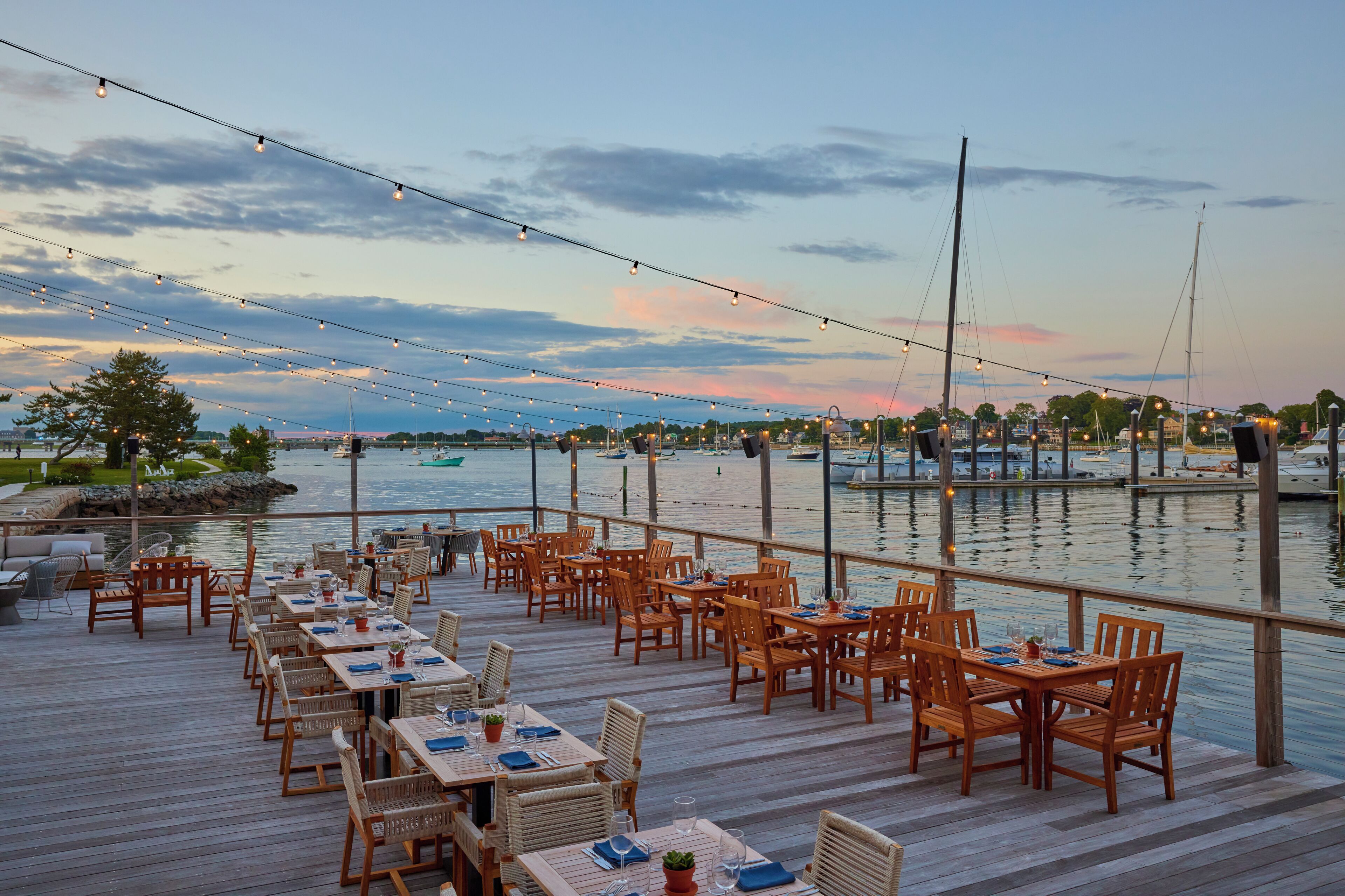 A waterfront restaurant with wooden tables and chairs, string lights, and a view of the marina.