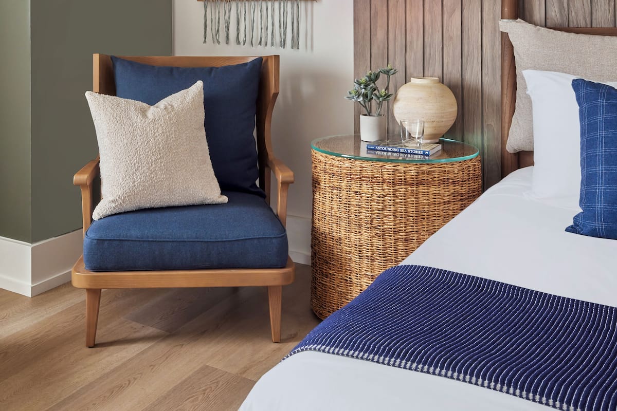 Un dormitorio con cabecera de madera, un sillón azul con almohada blanca, una mesita de noche con lámpara, un jarrón y un libro.