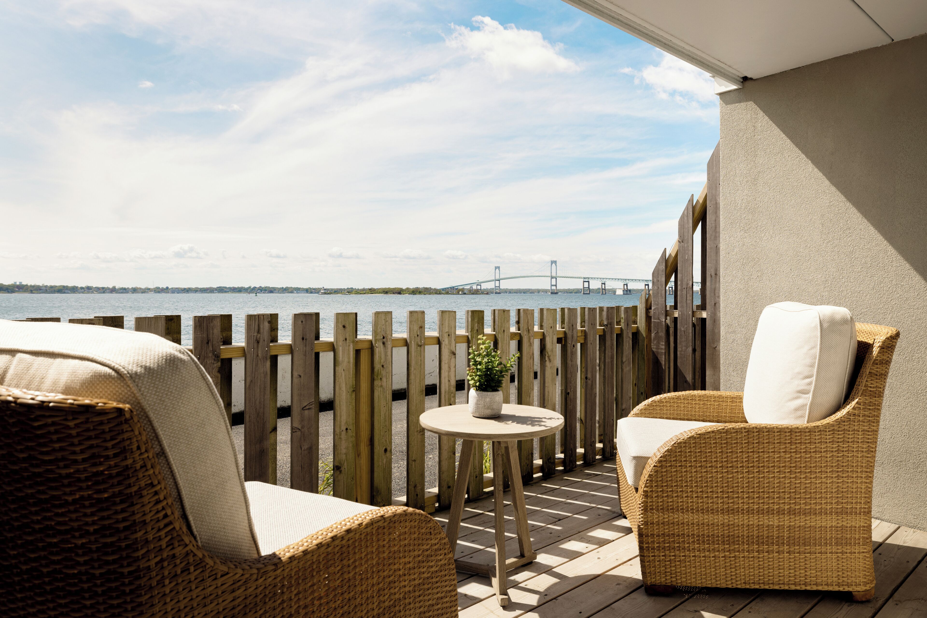 Ein Balkon mit Korbmöbeln, einem kleinen runden Tisch mit einer Pflanze und Blick auf eine Brücke über dem Wasser.
