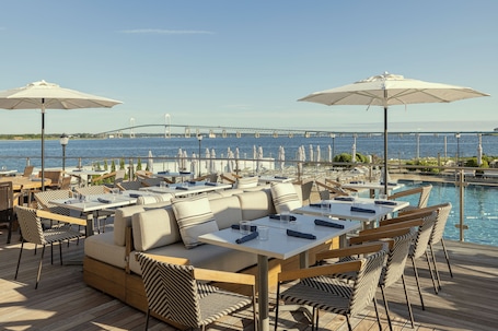 An outdoor dining area with tables and chairs, overlooking a body of water and a bridge.. Newport Harbor Island Resort