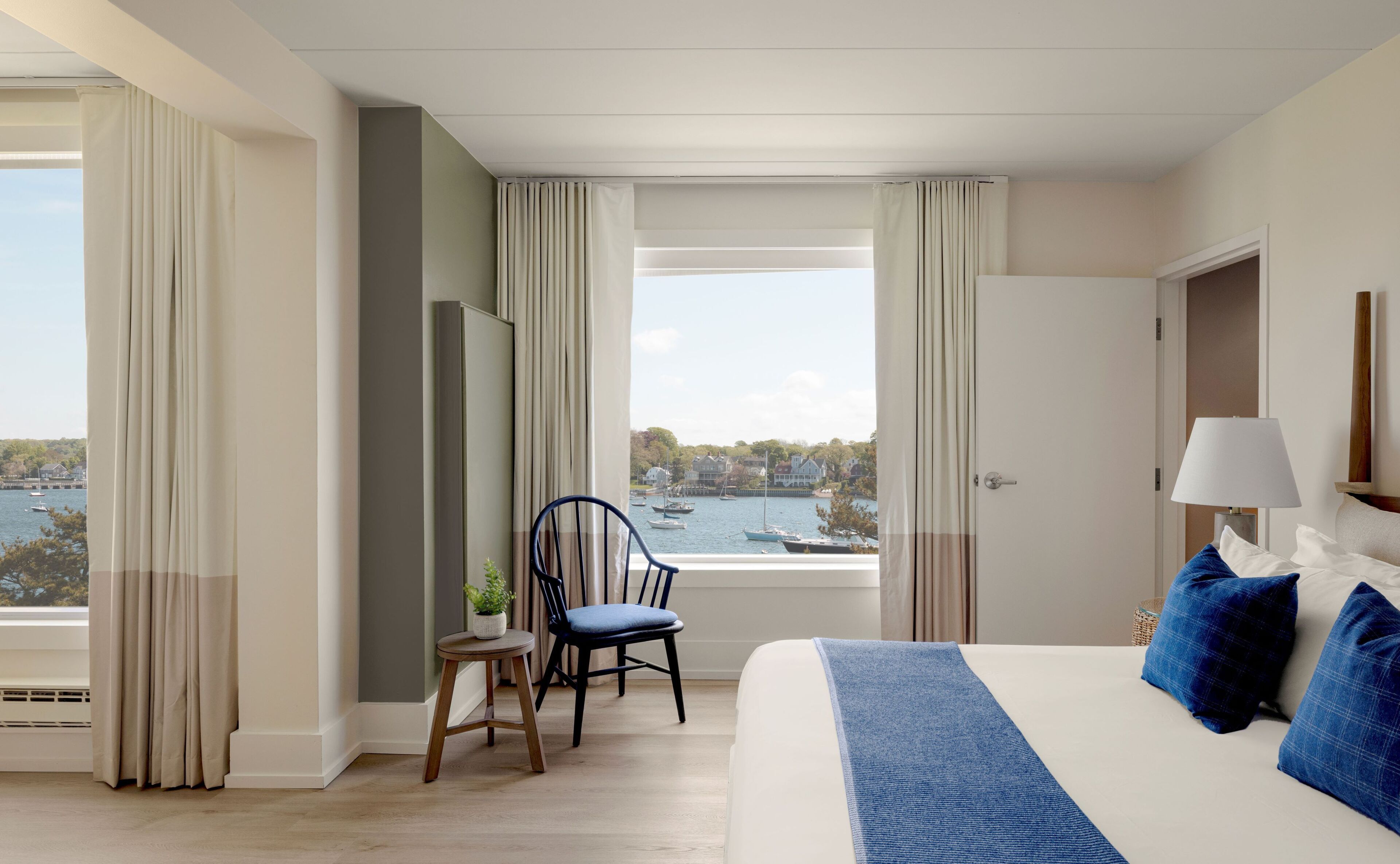 Une chambre d’hôtel avec un grand lit, une chaise, une petite table avec une plante, et une vue sur les bateaux et les maisons par la fenêtre.