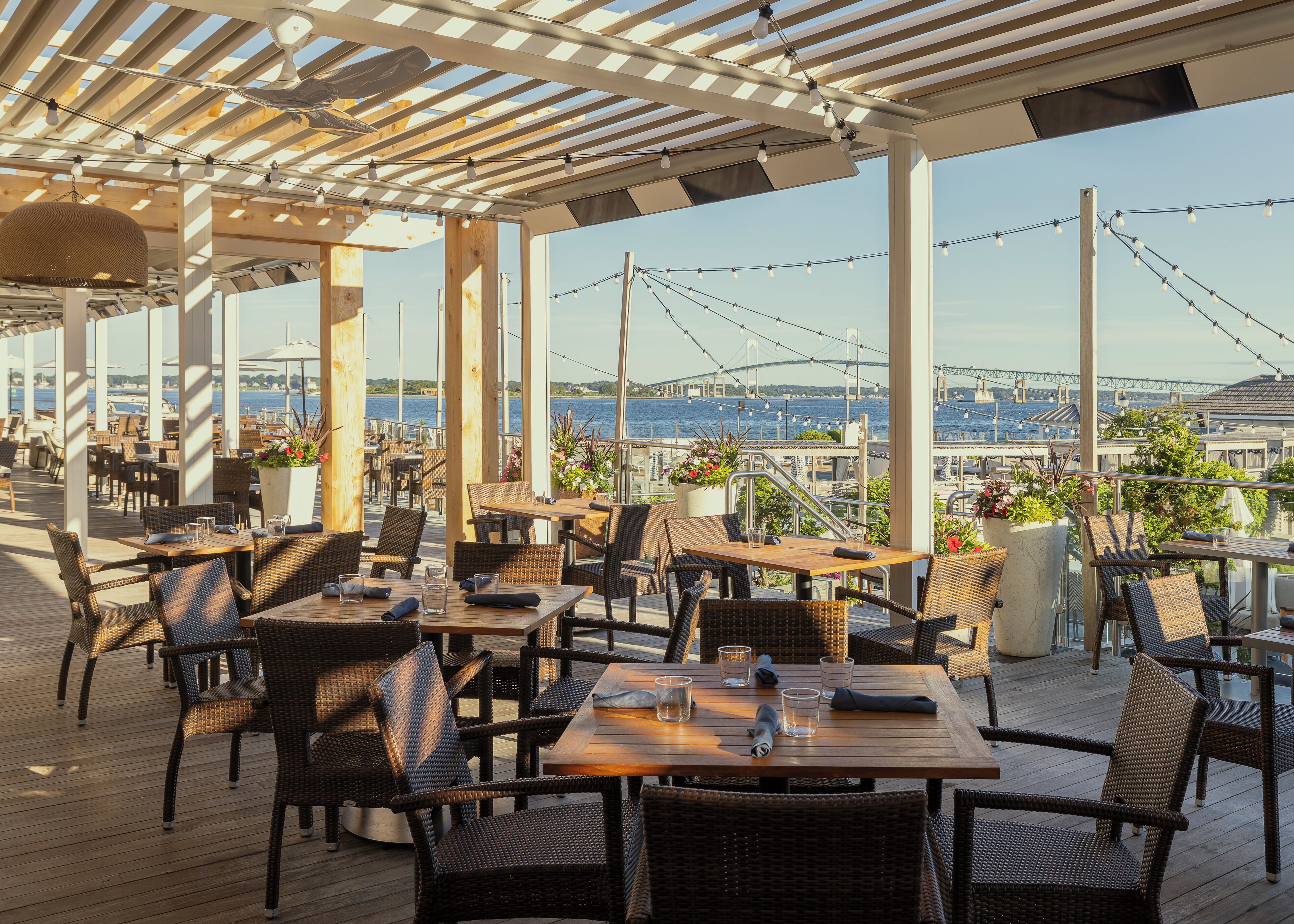 An outdoor dining area with wicker furniture and potted plants, overlooking a body of water and a bridge.