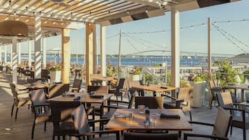An outdoor dining area with wicker furniture and potted plants, overlooking a body of water and a bridge.