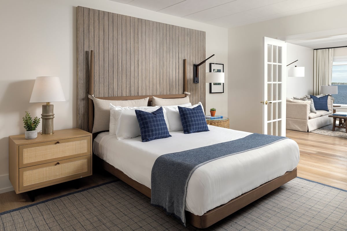 Un dormitorio moderno con una cama grande, una mesita de noche con lámpara y vistas al mar a través de una puerta abierta.