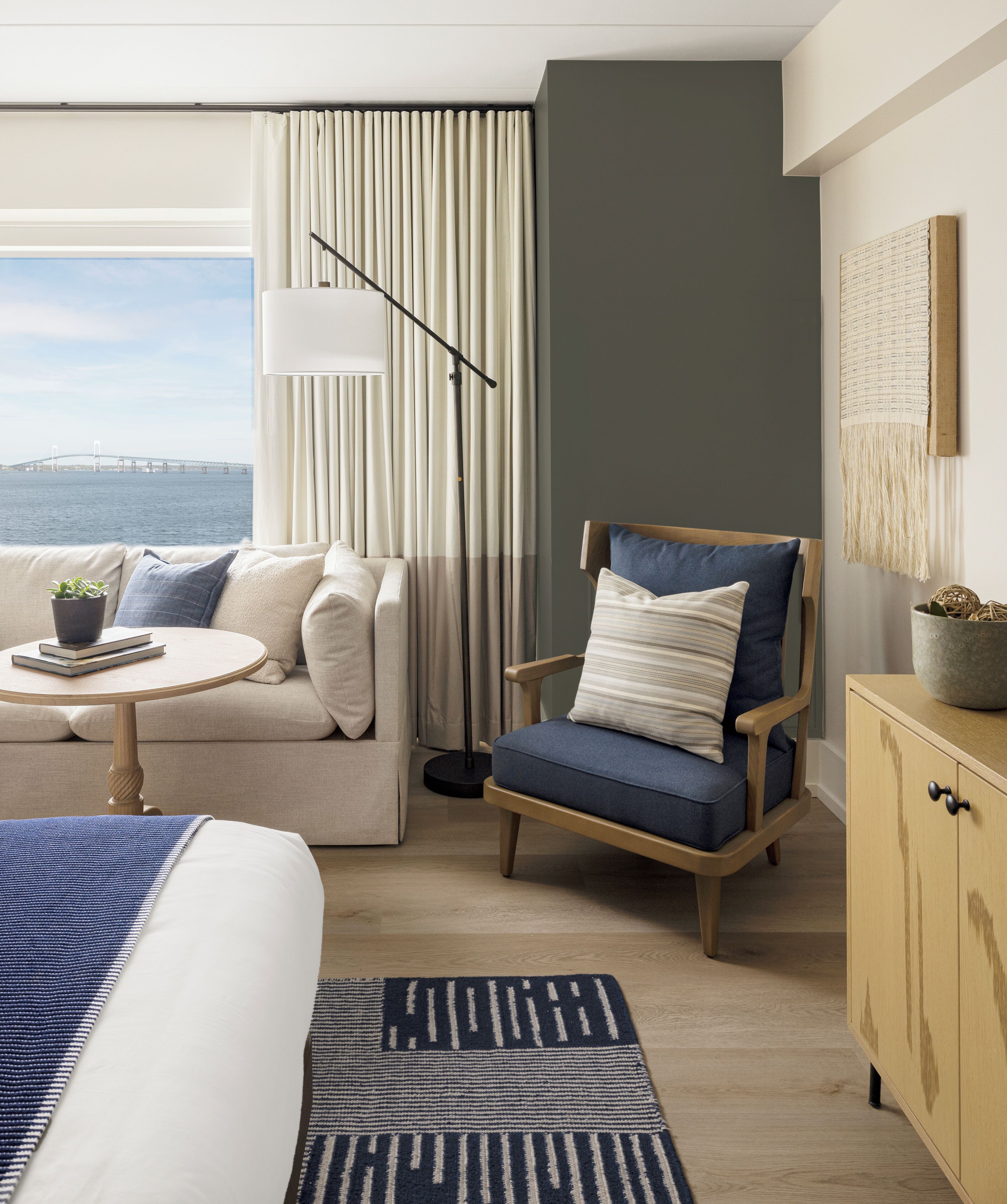 Habitación de hotel moderna con cama, sofá, sillón, mesita y vistas a un puente sobre el agua.