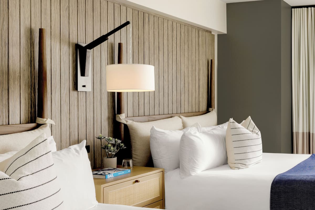 Habitación de hotel con dos camas, cabecero de madera, lámpara de noche y una mesita con un libro y una planta.