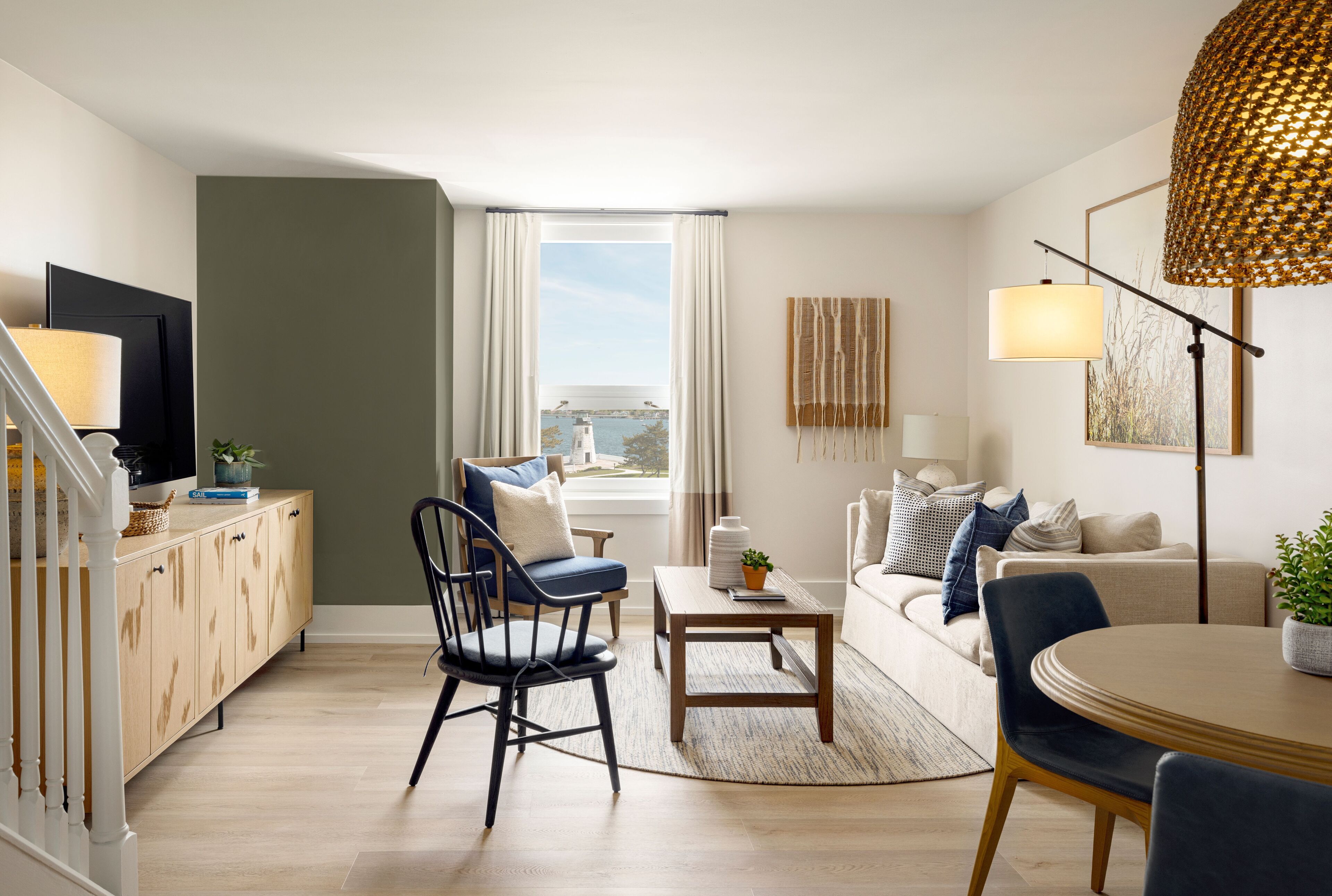 Una sala moderna con sofá, sillas, una mesa de centro y una mesa de comedor. Hay una ventana con cortinas, un televisor sobre un mueble bajo y una lámpara sobre un pedestal.