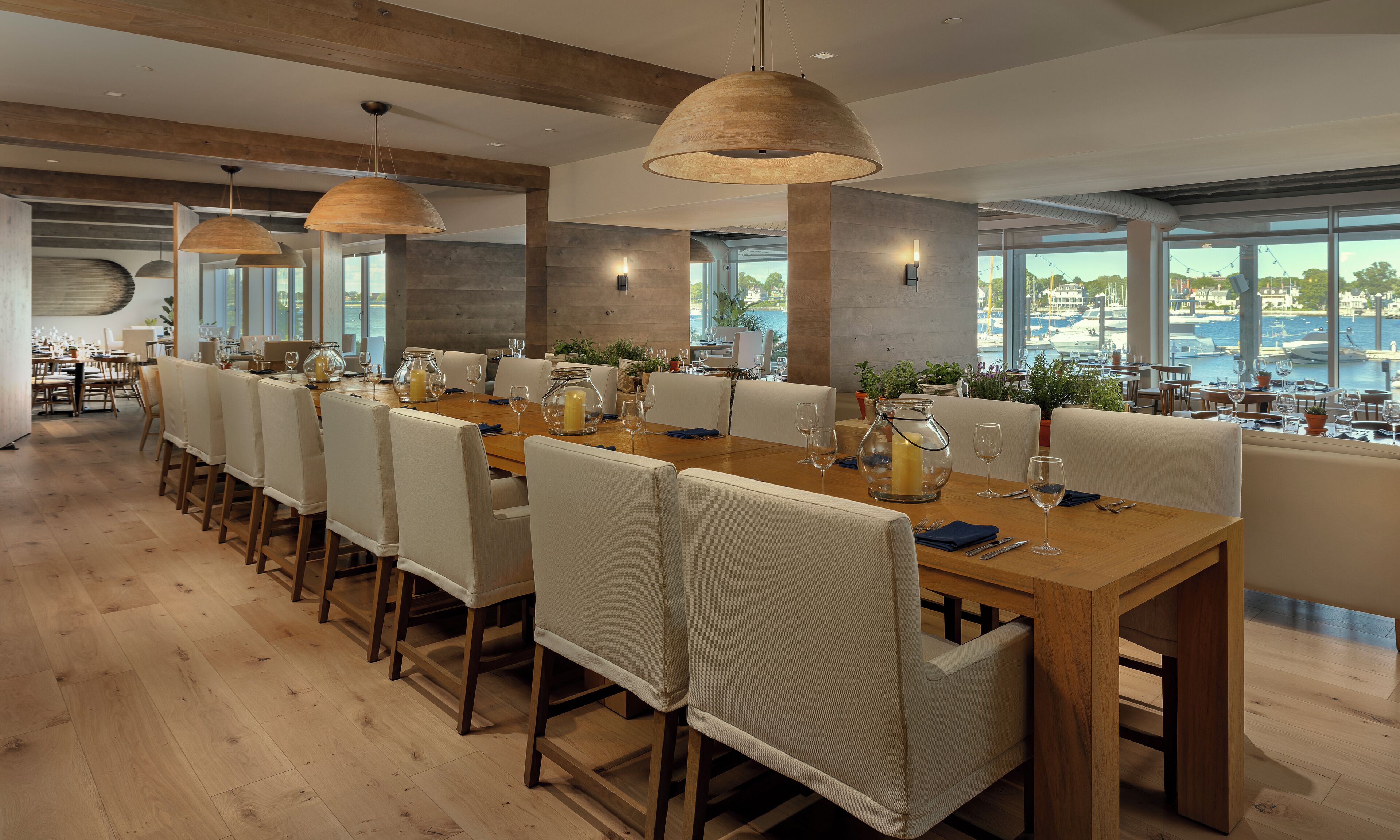 A modern dining area with long tables, white cushioned chairs, and large windows offering a view of the marina.
