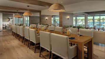 A modern dining area with long tables, white cushioned chairs, and large windows offering a view of the marina.