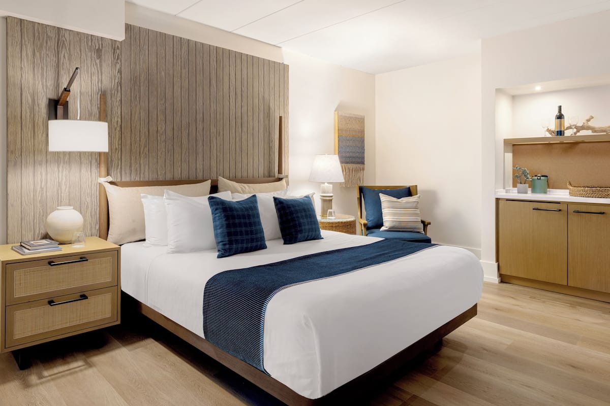 Habitación de hotel con una cama grande, cabecero de madera, mesitas de noche, un escritorio pequeño y una kitchenette.