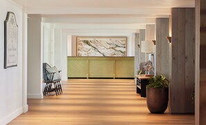 A hallway with wooden flooring, a green upholstered bench, a framed artwork on the wall, and a potted plant.