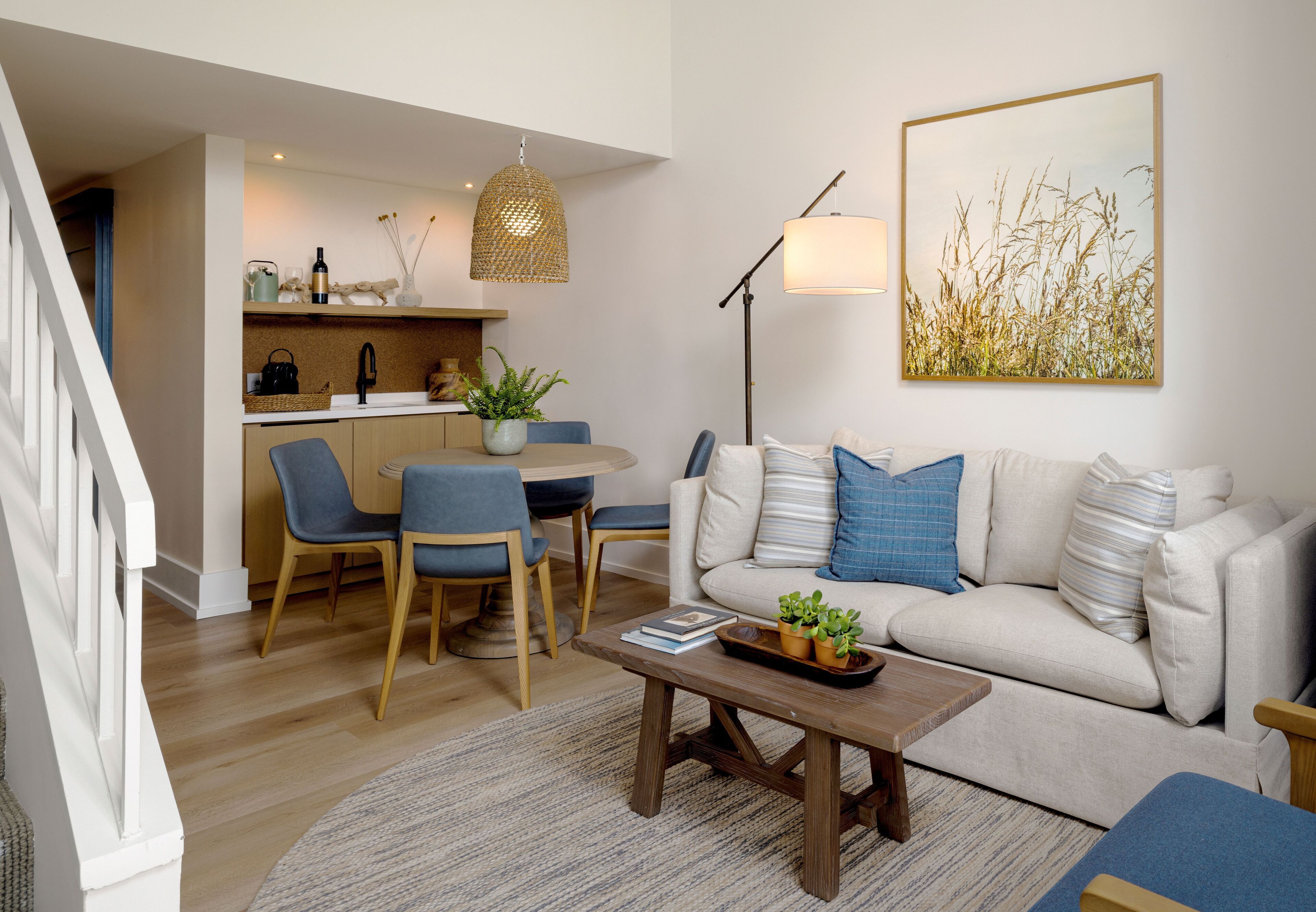 A modern living room with a sofa, coffee table, and chairs. There is a dining area with a table and chairs. A kitchen is visible in the background.