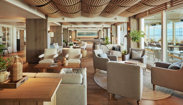 Amplia sala de estar con diversas opciones de asientos, techo de madera y ventanas grandes que ofrecen vista a un puente y al agua.