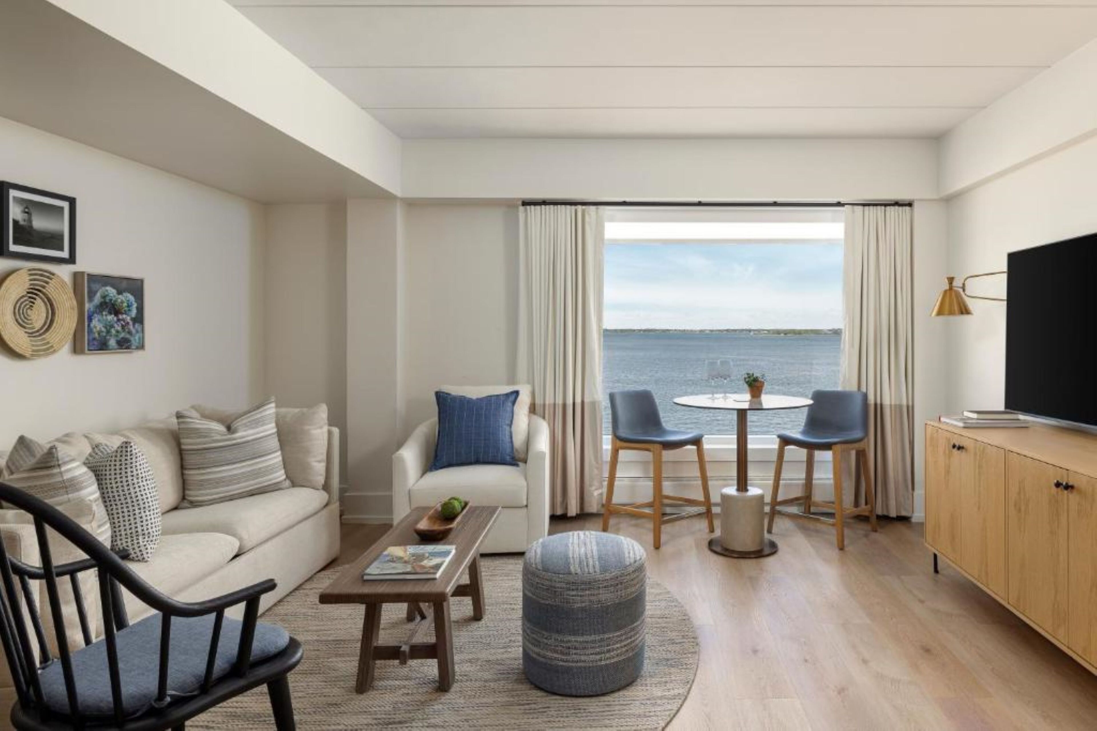 A modern living room with a sofa, chairs, a coffee table, and a TV. There is a view of the sea through large windows.