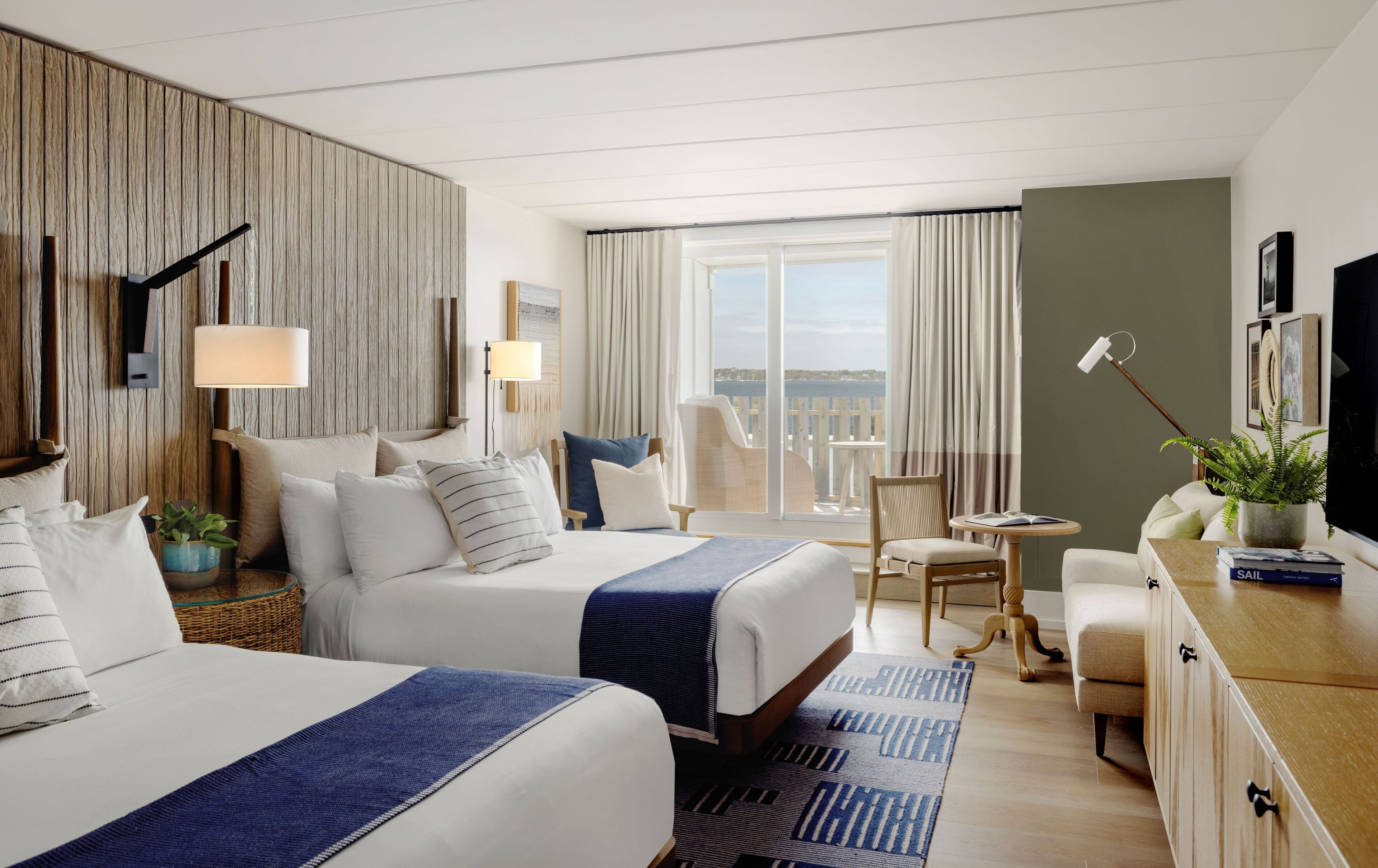 Ein Hotelzimmer mit zwei Betten, einem Balkon, einem Schreibtisch mit Pflanze und einem Fernseher.